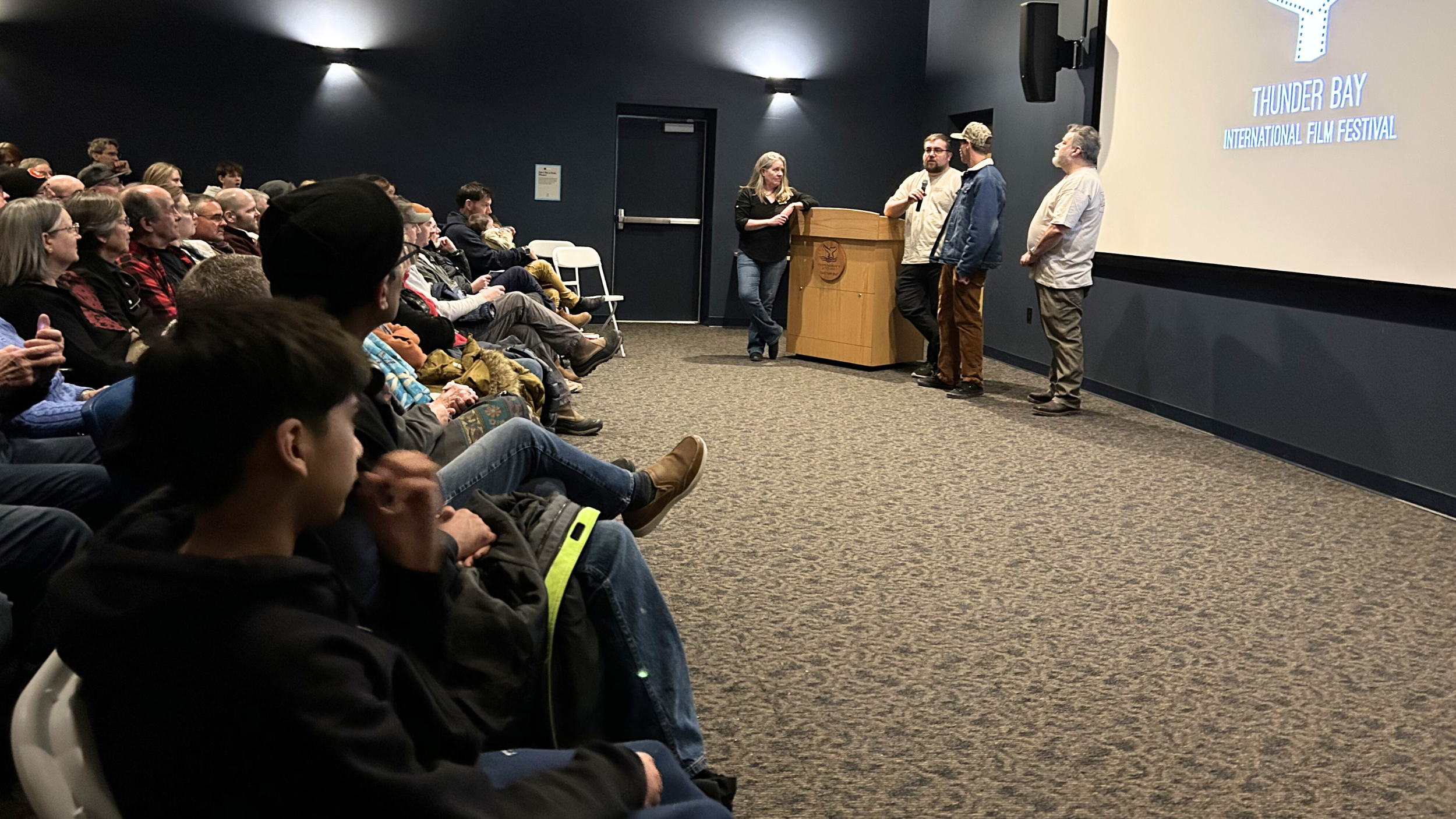 An auditorium with all the chairs filled by the audience and three men and one woman presenting at the front of the room.